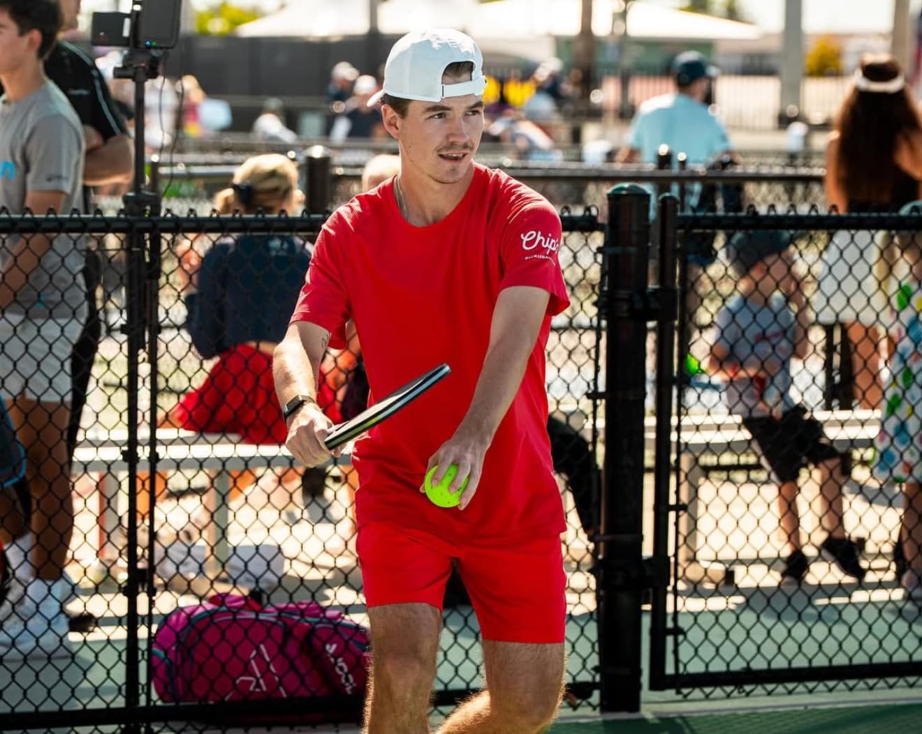 Adam Harvey in a red Chicken N Pickle shirt and white cap, paddle ready, on a sunlit tournament court
