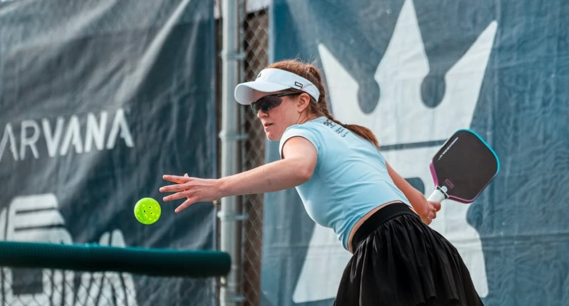 Liz Truluck reaching for a ball mid-rally, light blue top and white visor, NIRVANA tournament backdrop