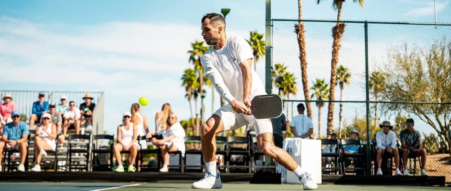 Mota Alhouni mid-backhand stretch in a white JOOLA kit, palm trees and sunlit crowd behind