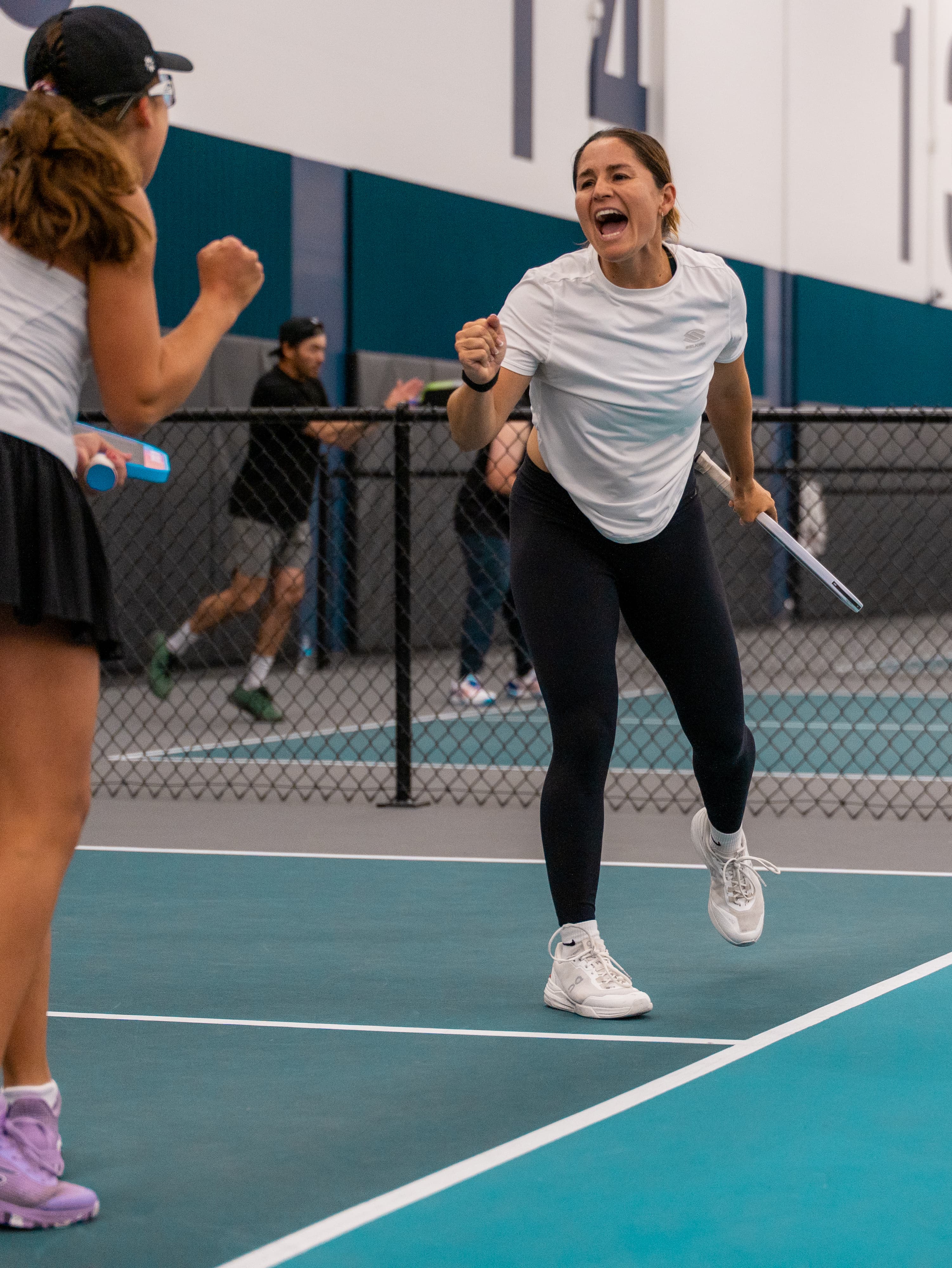 Charlie Treviño celebrating a point with a clenched fist, her doubles partner in the foreground at an indoor tournament