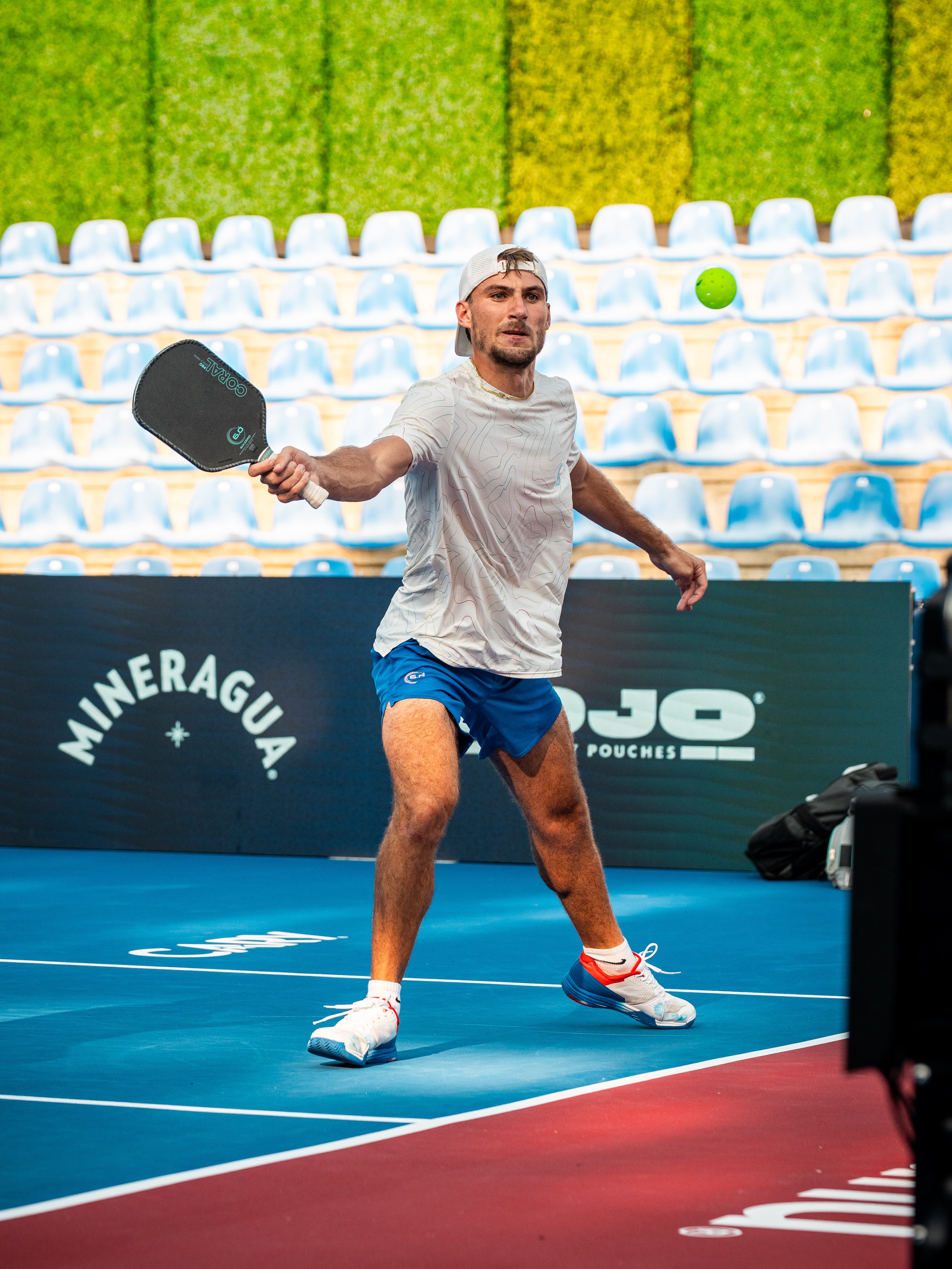 Zach Gonnering driving a forehand, ball in the air beside him, empty stadium seating behind the court