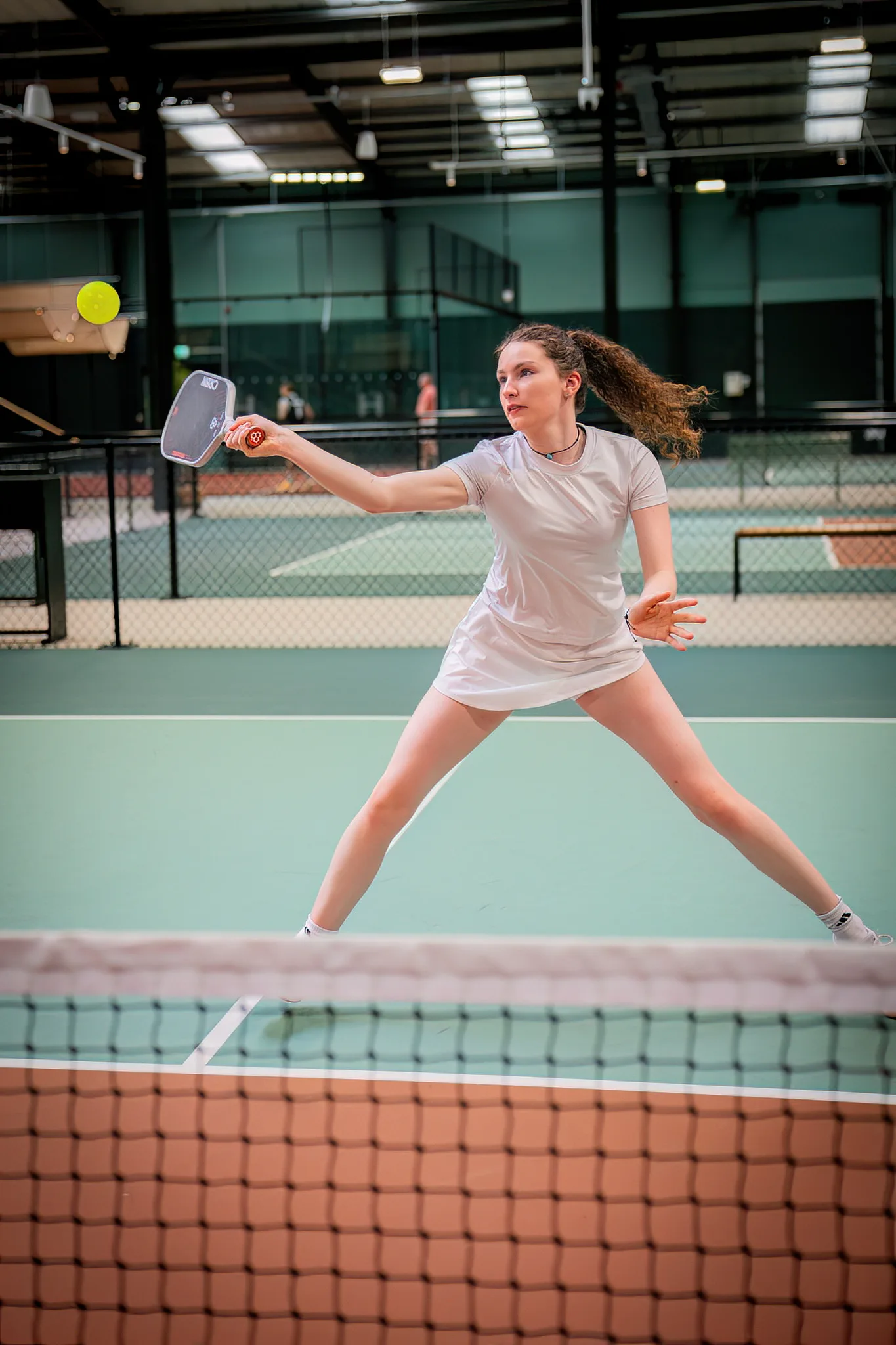 Ellie Tomkinson stretching wide for a forehand volley at the net on an indoor court